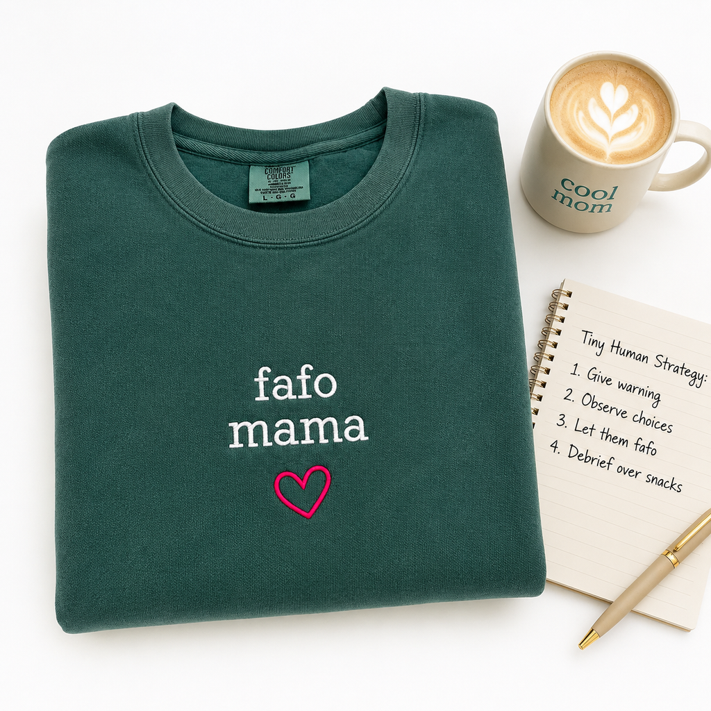 A folded dark green crewneck sweatshirt with 'fafo mama' and a pink heart embroidered on the front, styled in a flat lay with a coffee mug, a notebook, and a pen on a white background.