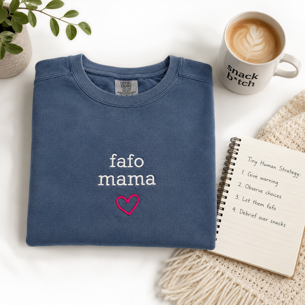 A folded slate blue crewneck sweatshirt with 'fafo mama' and a pink heart embroidered on the chest, styled in a cozy flat-lay arrangement with a coffee mug, notebook, and plant.