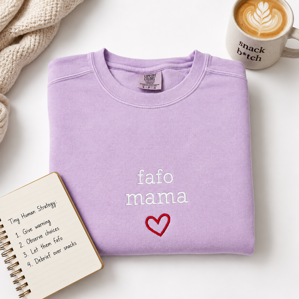 A folded light purple sweatshirt with 'fafo mama' and a dark pink heart embroidered on the front, styled on a white surface with a mug, notebook, and knit blanket.