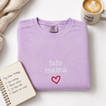 A folded light purple sweatshirt with 'fafo mama' and a dark pink heart embroidered on the front, styled on a white surface with a mug, notebook, and knit blanket.