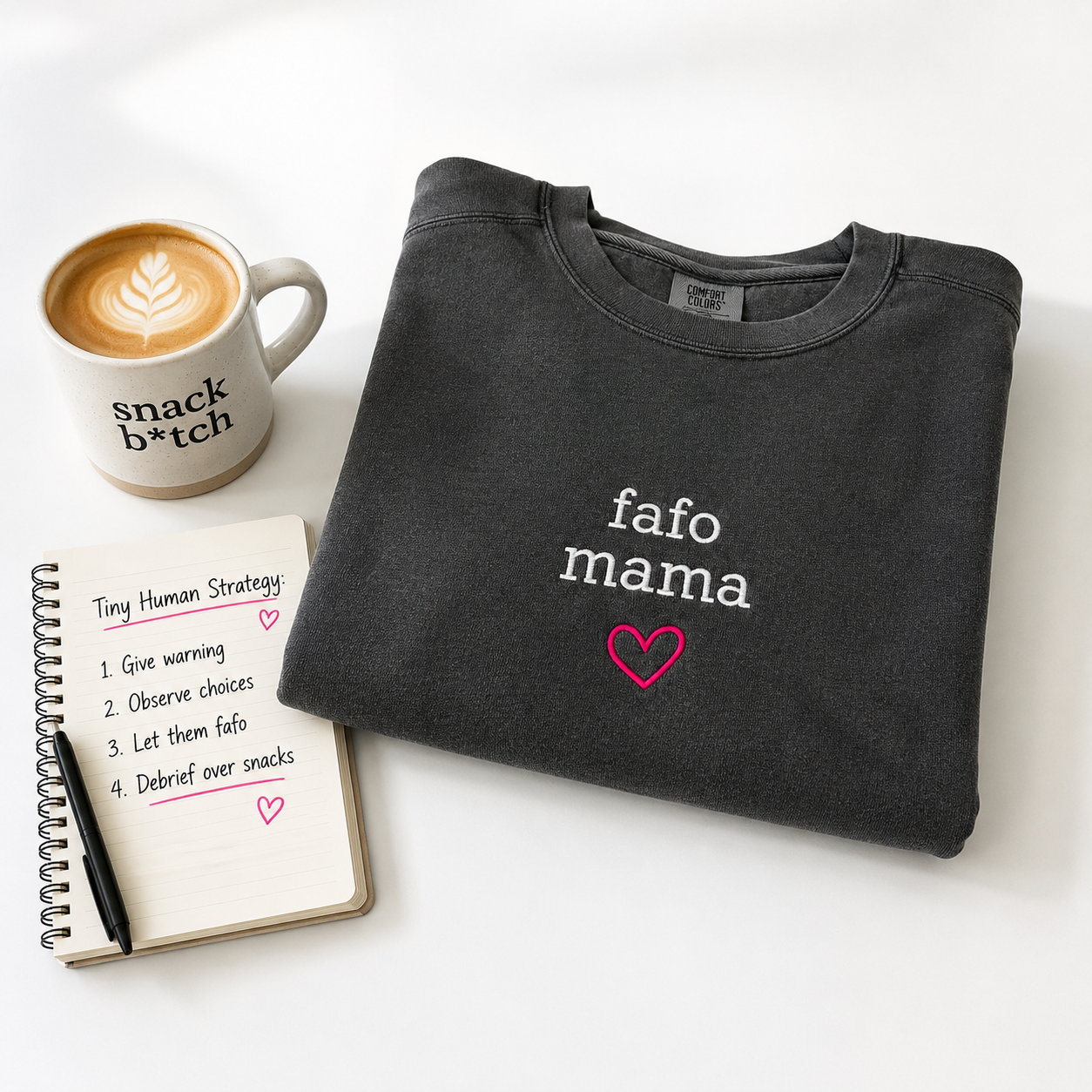 A folded dark charcoal crewneck sweatshirt with the words 'fafo mama' in white and a pink heart embroidered, styled on a white background with a coffee mug, notebook, and pen.