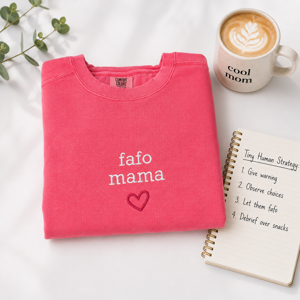 A folded bright pink sweatshirt with the words 'fafo mama' and a small heart outline embroidered in white and dark pink, styled on a white surface with a coffee mug, a notebook, and green leaves.
