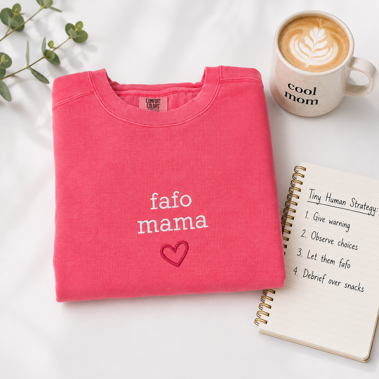 A folded bright pink sweatshirt with the words 'fafo mama' and a small heart outline embroidered in white and dark pink, styled on a white surface with a coffee mug, a notebook, and green leaves.