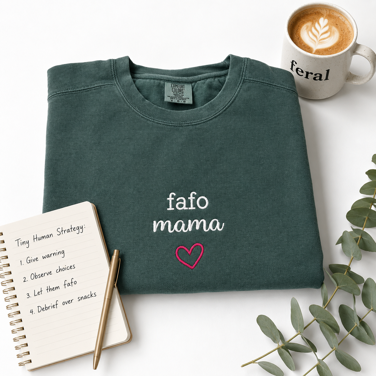 A folded dark teal sweatshirt with 'fafo mama' and a pink heart embroidered on the front, styled in a flat lay with a coffee mug, notebook, pen, and eucalyptus leaves on a white background.