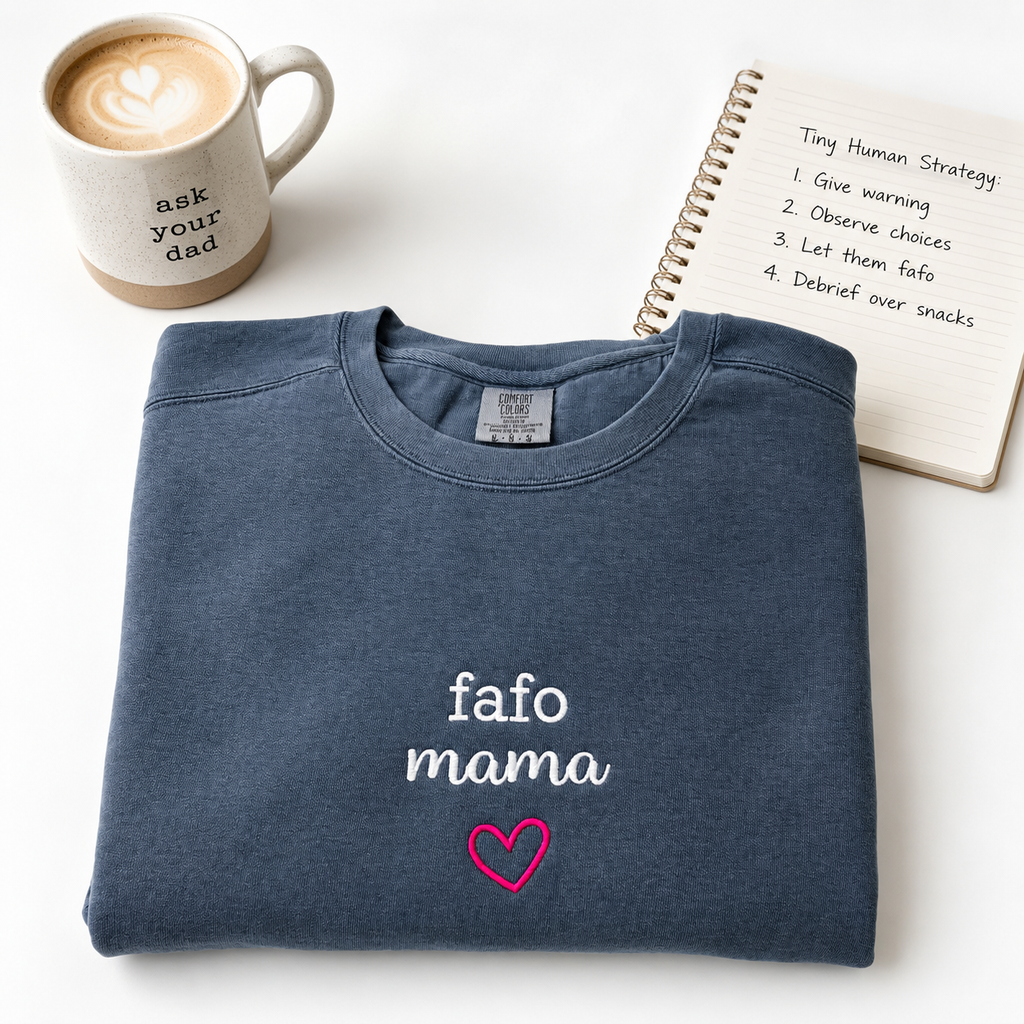 A folded navy blue crewneck sweatshirt with 'fafo mama' embroidered in white and a pink heart, styled on a white background with a coffee mug and a notebook.