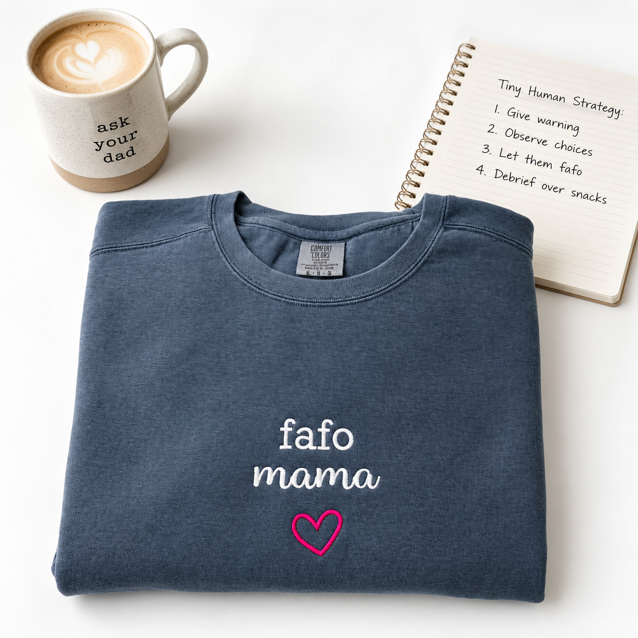 A folded navy blue crewneck sweatshirt with 'fafo mama' embroidered in white and a pink heart, styled on a white background with a coffee mug and a notebook.
