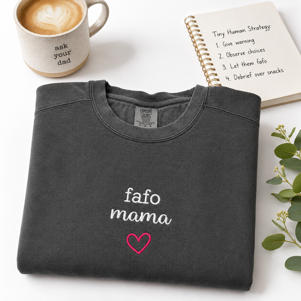 A folded dark gray crewneck sweatshirt with 'fafo mama' and a pink heart embroidered on the front, styled on a white surface with a coffee mug, a notebook, and greenery.