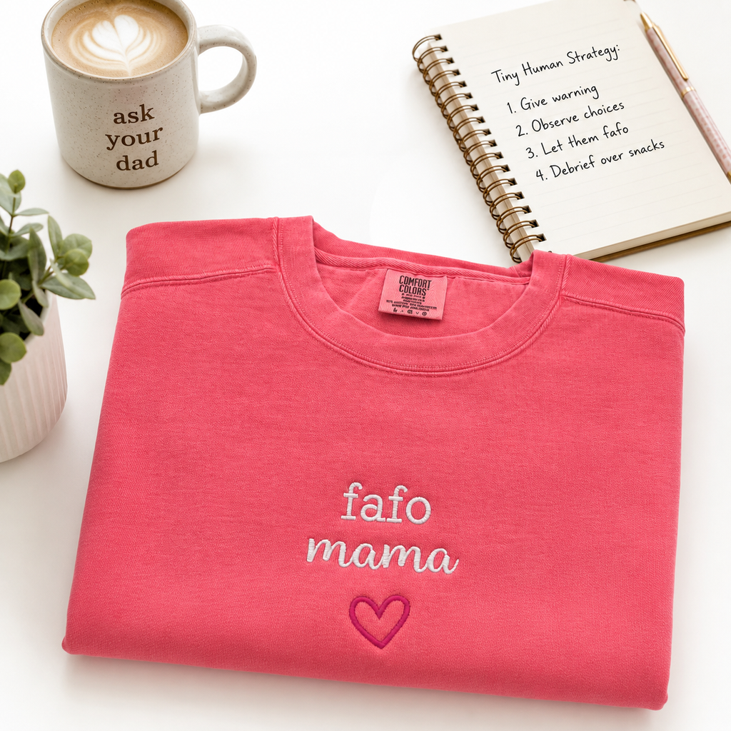 A folded coral pink crewneck sweatshirt with 'fafo mama' and a heart embroidered on the front, styled on a white surface with a coffee mug, notebook, and plant.