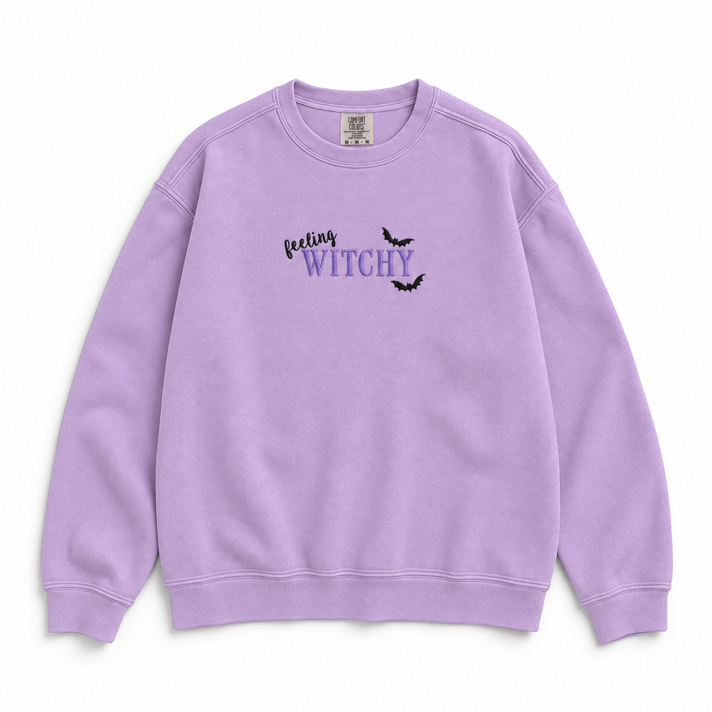 A lavender purple crewneck sweatshirt featuring the graphic text 'feeling WITCHY' in black and purple with two small black bats, displayed on a clean white background.