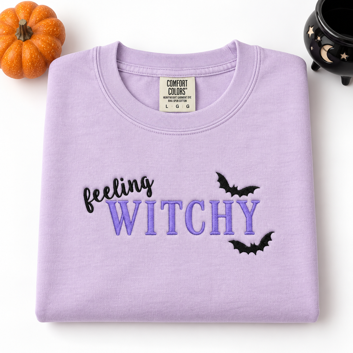 A folded lavender crewneck sweatshirt with 'feeling WITCHY' embroidered on the front, styled with a small orange pumpkin and a black cauldron on a white background.