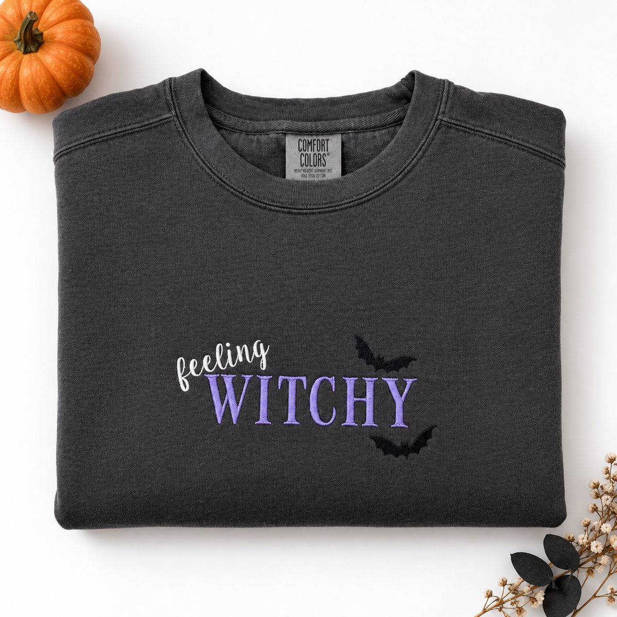 A folded dark charcoal gray sweatshirt on a white background, featuring the text 'feeling WITCHY' in white and lavender-purple across the chest, with two black bat silhouettes. A small orange pumpkin is in the top-left corner and dried flowers are in the bottom-right corner.