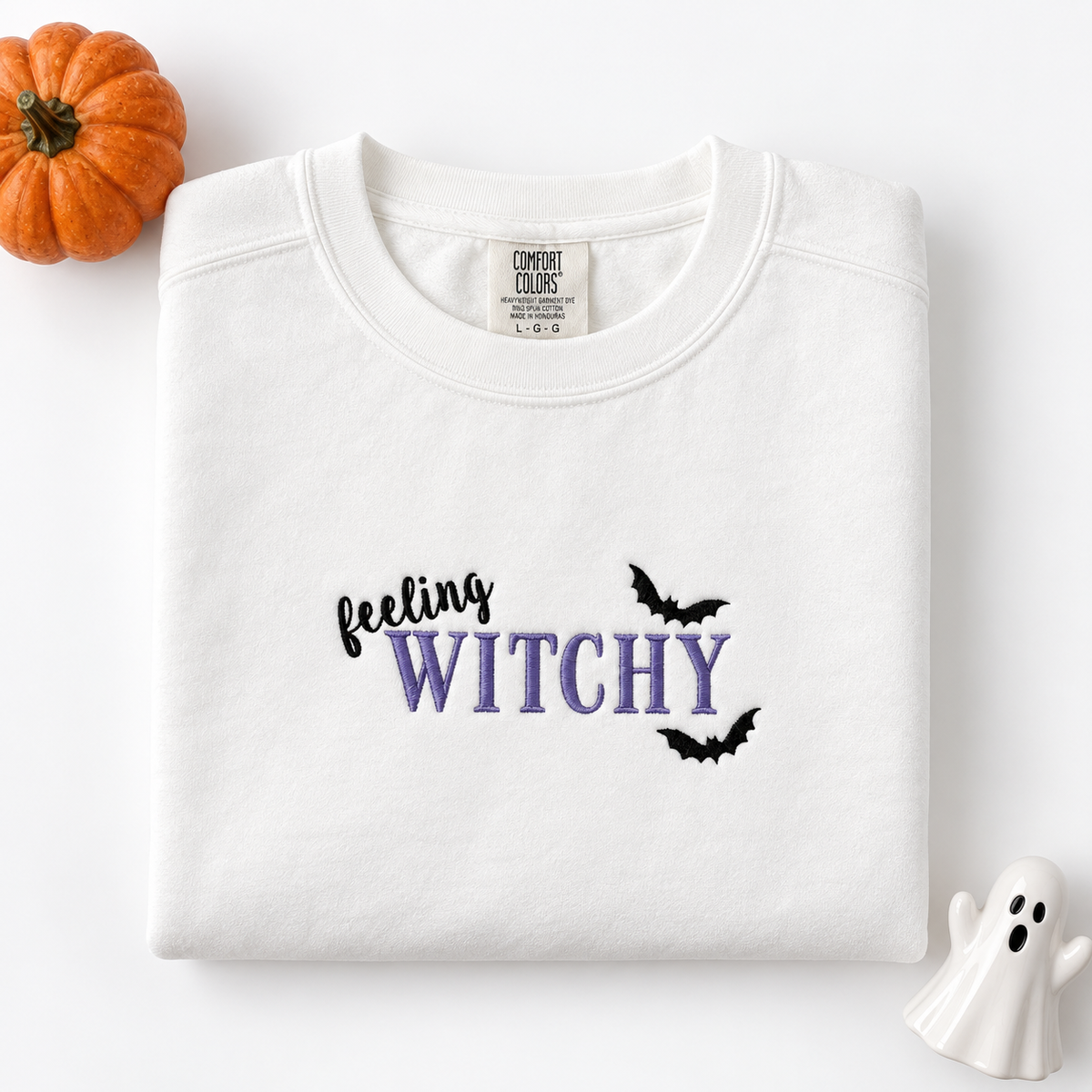 A folded white crewneck sweatshirt with 'feeling WITCHY' embroidered on the chest, styled with a small orange pumpkin and a white ghost decoration on a white background.
