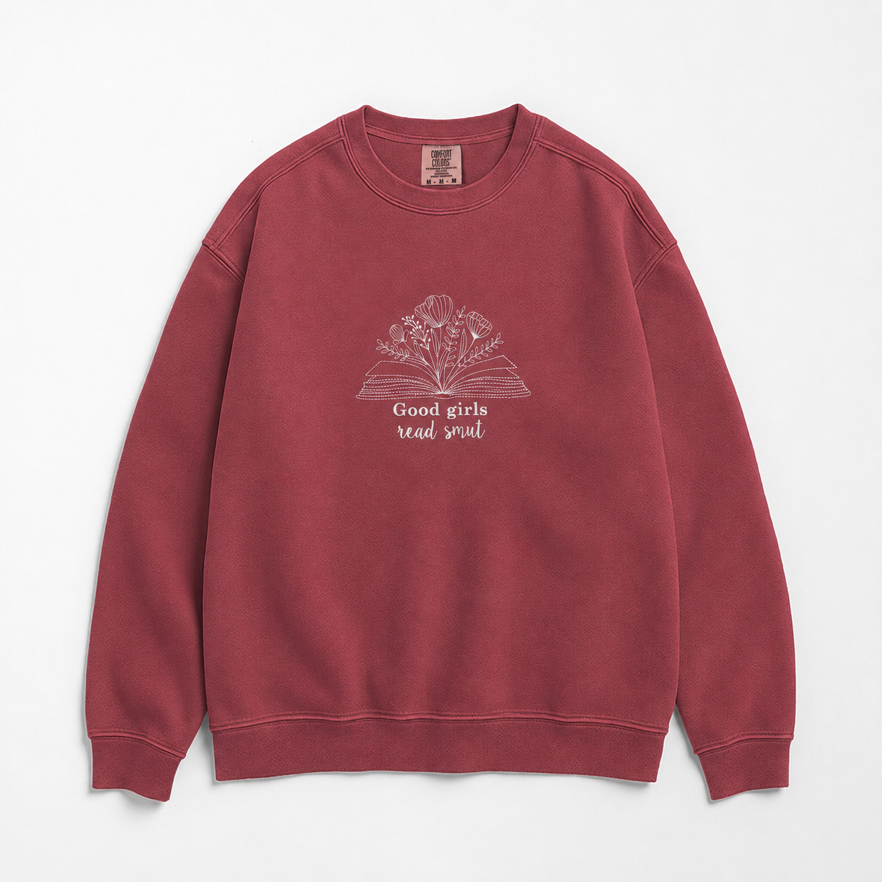 A crewneck sweatshirt in a washed, muted brick red color, featuring a graphic of an open book with flowers and the text 'Good girls read smut' on the chest, laid flat against a light gray background.
