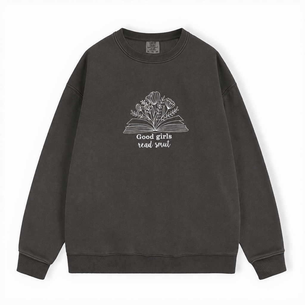 A dark gray washed crewneck sweatshirt featuring a white graphic of an open book with flowers and the text 'Good girls read smut' on the chest, displayed flat against a white background.