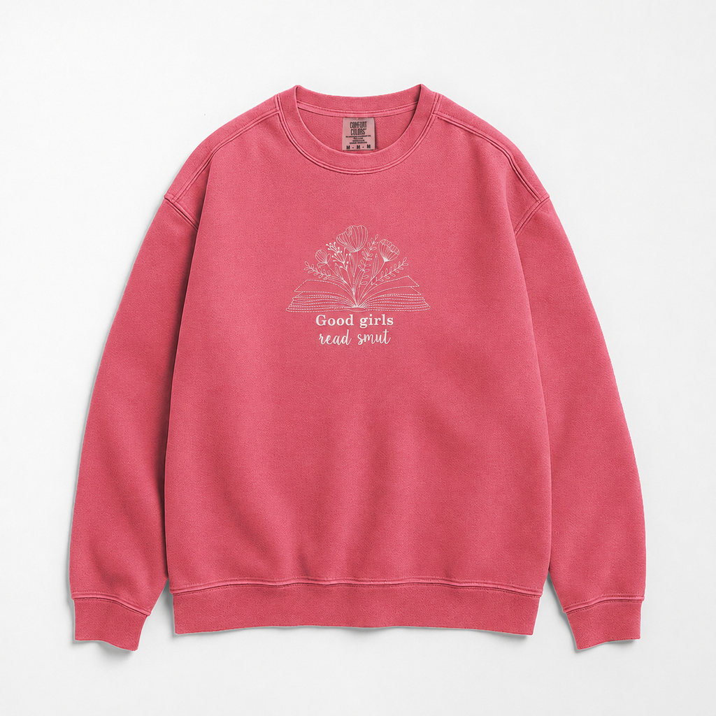 A pink crewneck sweatshirt featuring a white graphic of an open book with flowers and the text 'Good girls read smut' on the front, displayed against a clean white background.