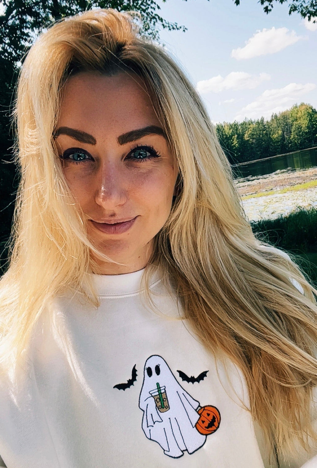 A woman with long blonde hair wearing a white crewneck sweatshirt with a cartoon ghost holding a pumpkin and bats, photographed outdoors with trees and water in the background.