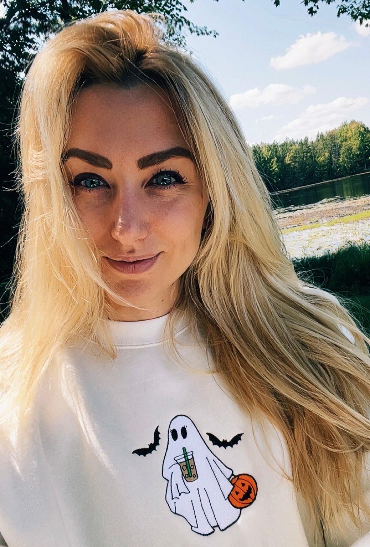 A woman with long blonde hair wearing a white crewneck sweatshirt with a cartoon ghost holding a pumpkin and bats, photographed outdoors with trees and water in the background.
