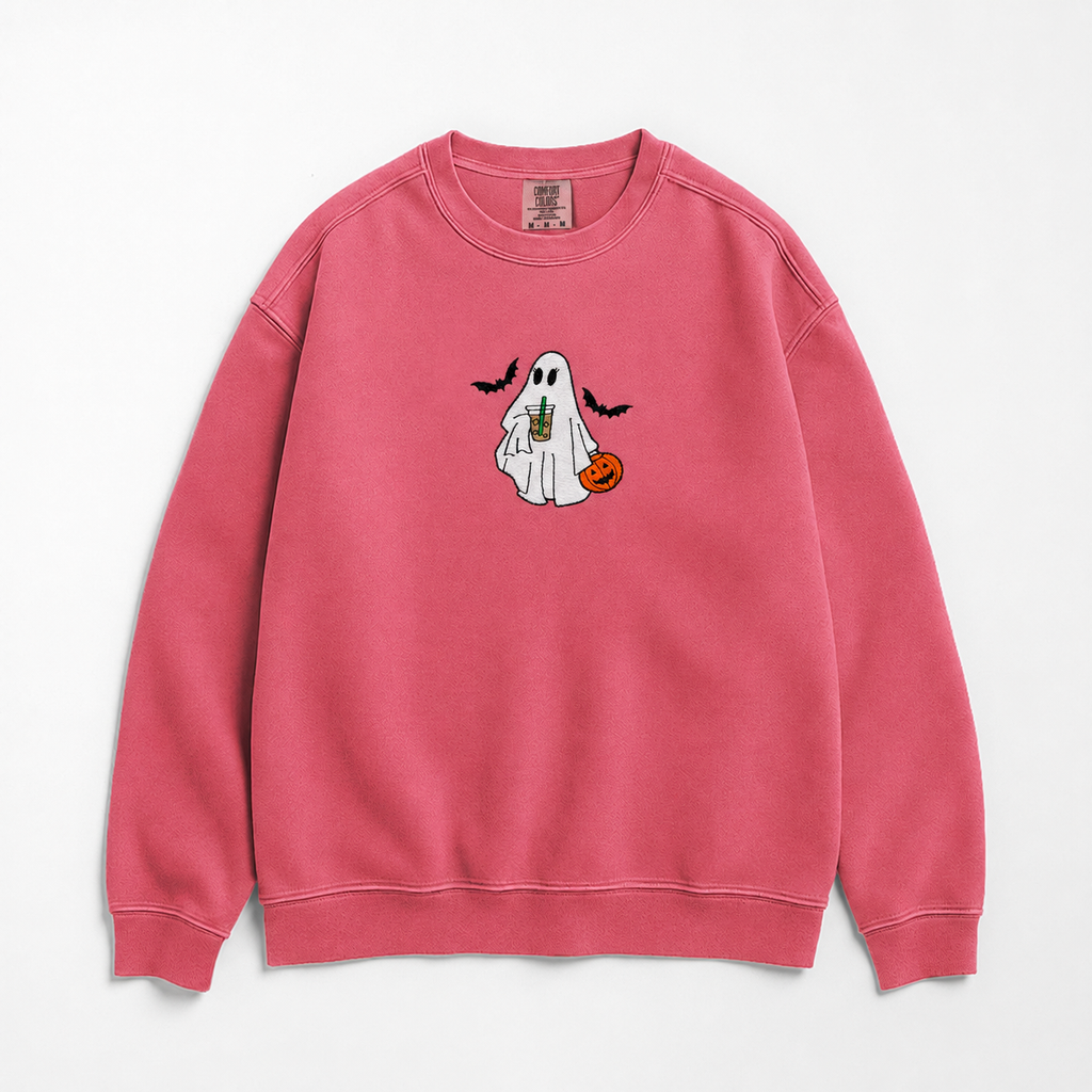 A pink crewneck sweatshirt featuring a cute cartoon ghost graphic on the chest, holding an iced coffee and a small orange pumpkin, with two black bats flying above. The sweatshirt is laid flat against a clean white background.