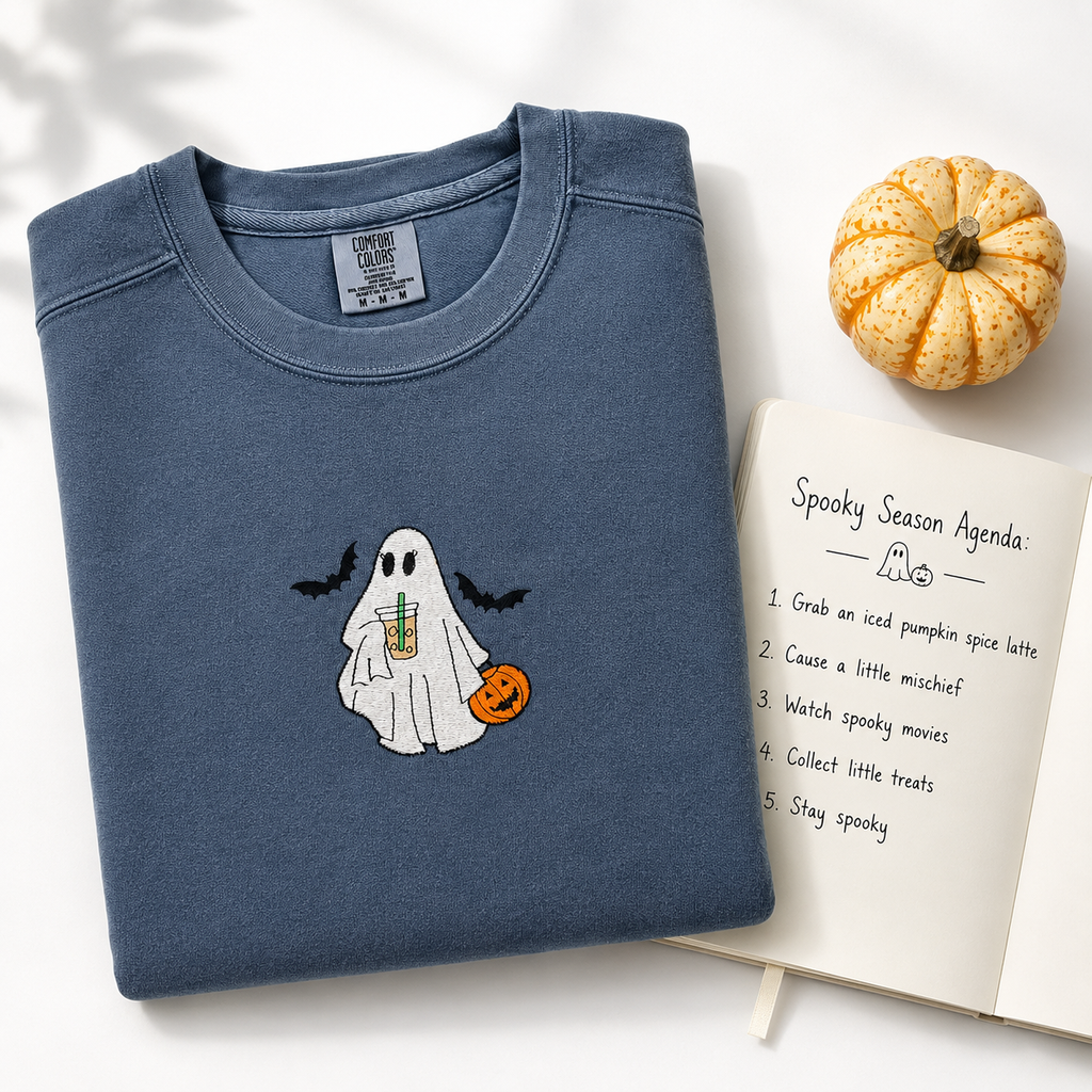 A folded slate blue crewneck sweatshirt with a cute embroidered ghost holding an iced coffee drink and a pumpkin, styled on a white background with a small pumpkin and an open notebook.