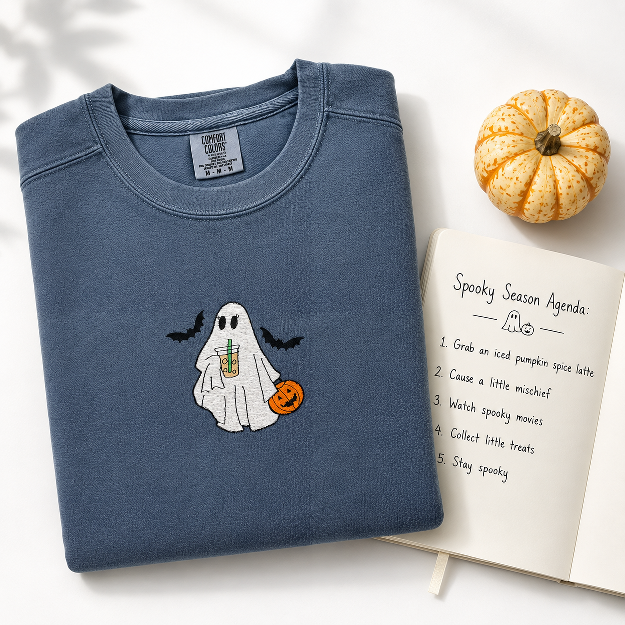 A folded slate blue crewneck sweatshirt with a cute embroidered ghost holding an iced coffee drink and a pumpkin, styled on a white background with a small pumpkin and an open notebook.