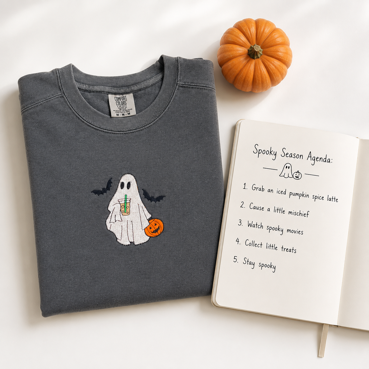 A folded dark gray crewneck sweatshirt with a cute embroidered ghost holding a pumpkin and an iced coffee drink, styled next to an open notebook with a 'Spooky Season Agenda' and a small orange pumpkin on a white background.