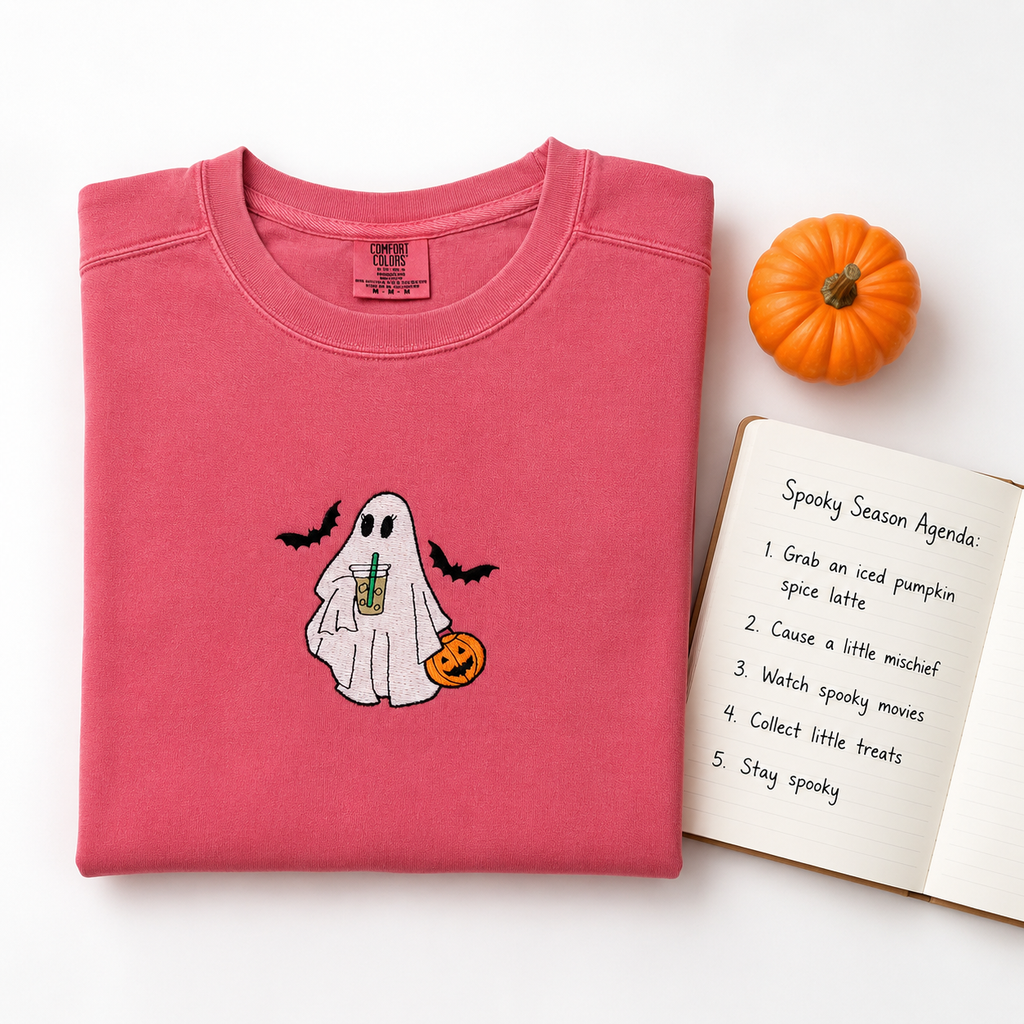 A folded pink crewneck sweatshirt with  an embroidered cute ghost graphic holding a pumpkin and an iced latte, styled on a white background with a small orange pumpkin and an open notebook listing spooky season agendas.