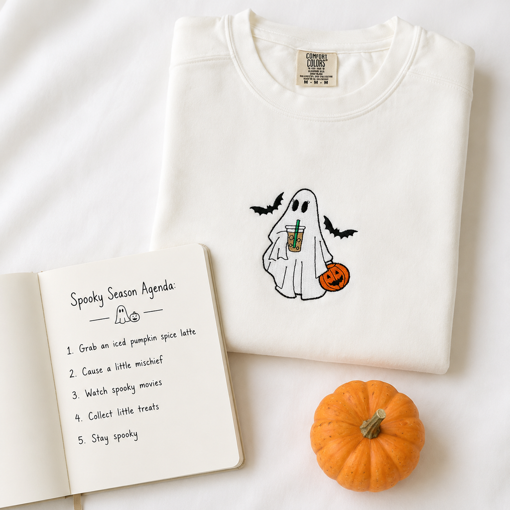 A folded white crewneck sweatshirt with an embroidered cute ghost graphic holding an iced coffee drink and a pumpkin, styled on a white background with an open 'Spooky Season Agenda' notebook and a small orange pumpkin.