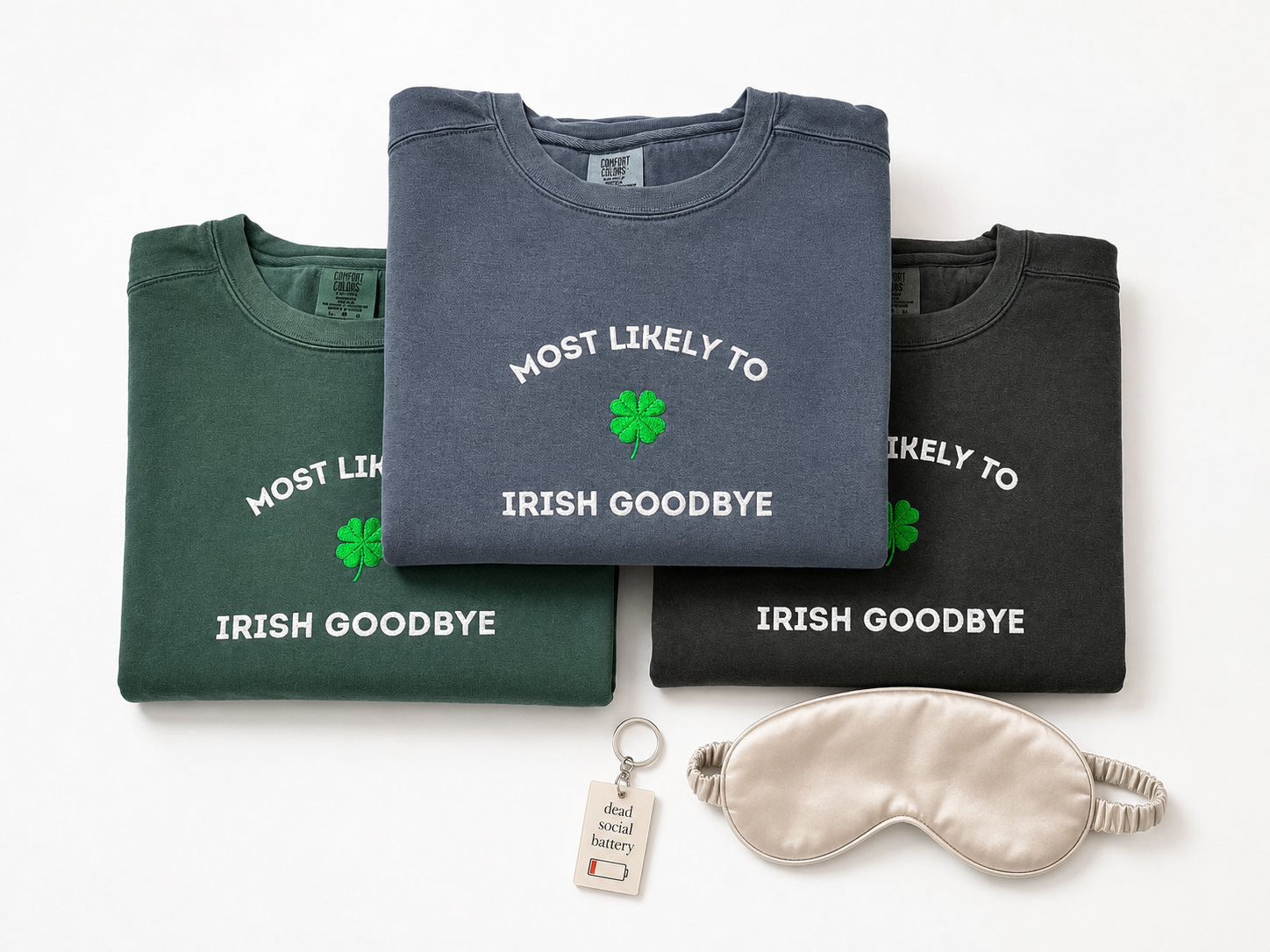 Three folded crewneck sweatshirts in green, slate blue, and charcoal gray, each printed with the phrase 'MOST LIKELY TO IRISH GOODBYE' and a green shamrock. A cream sleep mask and a small keychain tag are arranged in front on a white background.