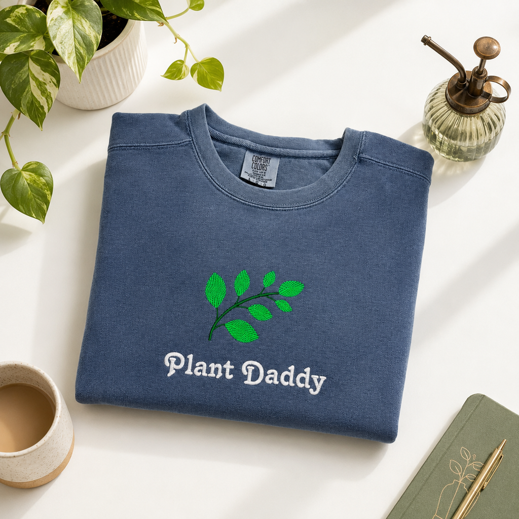 A folded navy blue crewneck sweatshirt with an embroidered green leaf graphic and the words 'Plant Daddy' in white, styled on a white surface with a plant, coffee cup, and notebook.