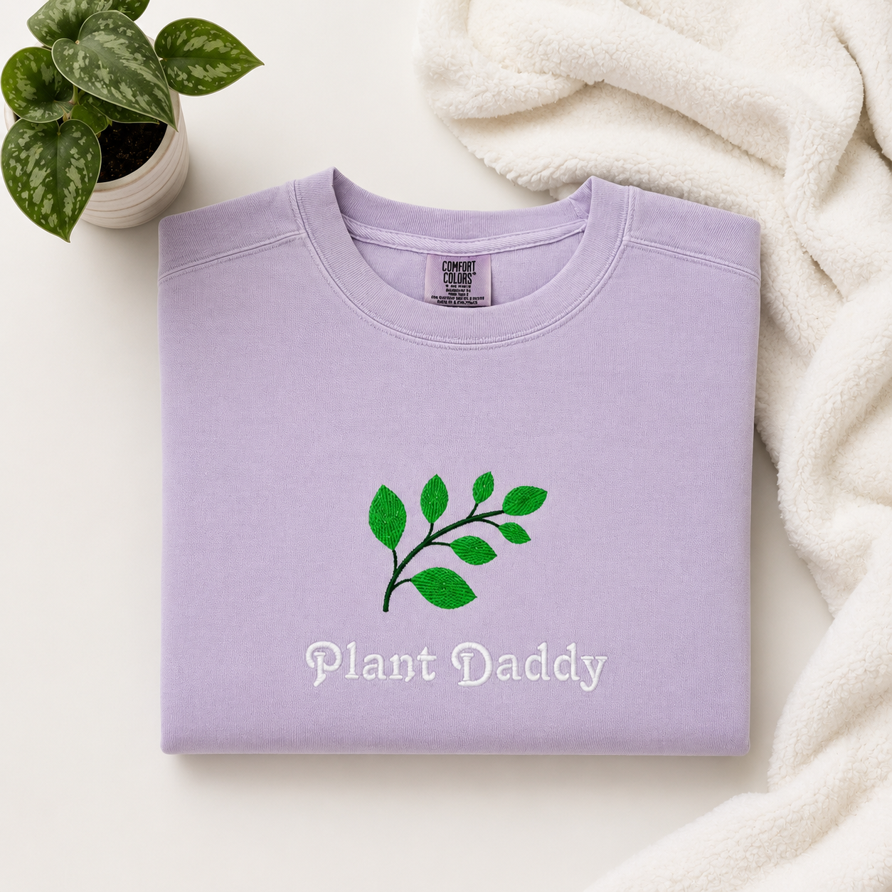 A folded light purple sweatshirt with an embroidered green leaf graphic and the words 'Plant Daddy' printed on the front, styled on a light surface with a small potted plant and a white textured blanket.