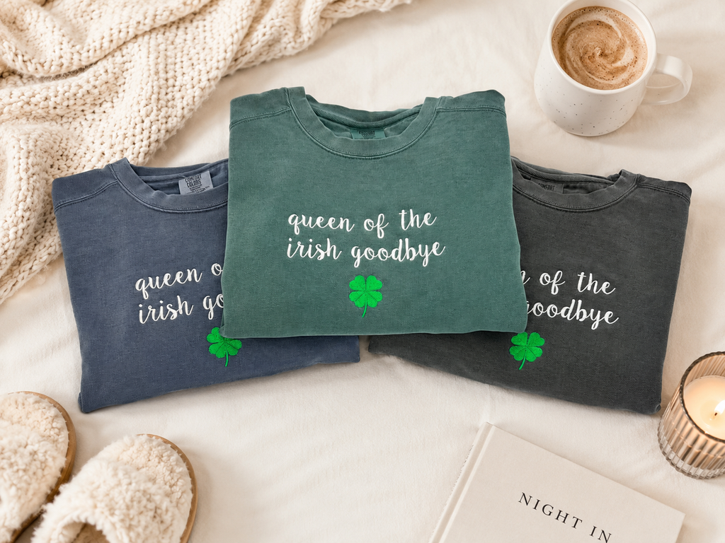 Three folded graphic t-shirts in slate blue, sage green, and charcoal gray are arranged in a cozy flat-lay with a knitted blanket, a cup of hot chocolate, slippers, and a candle. The center green shirt is the most prominent, featuring the white script text 'queen of the irish goodbye' and a green shamrock.