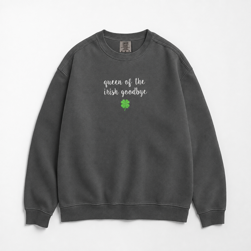 A dark gray crewneck sweatshirt featuring  embroidered text of 'queen of the irish goodbye' in white script and a small green four-leaf clover graphic on the chest, displayed flat against a light gray background.