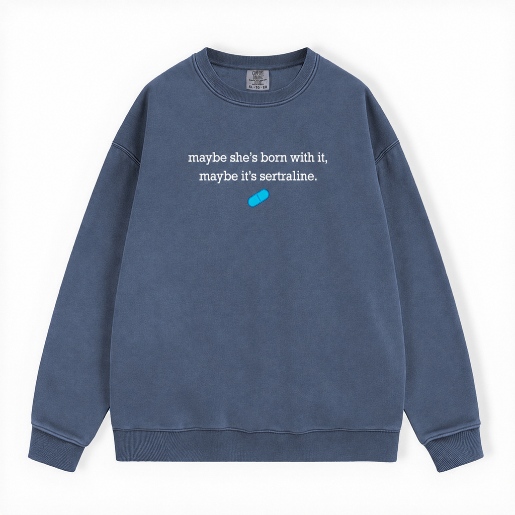 A washed slate blue crewneck sweatshirt featuring the white printed text 'maybe she's born with it, maybe it's sertraline.' and a small bright blue pill graphic on the chest, displayed flat against a white background.