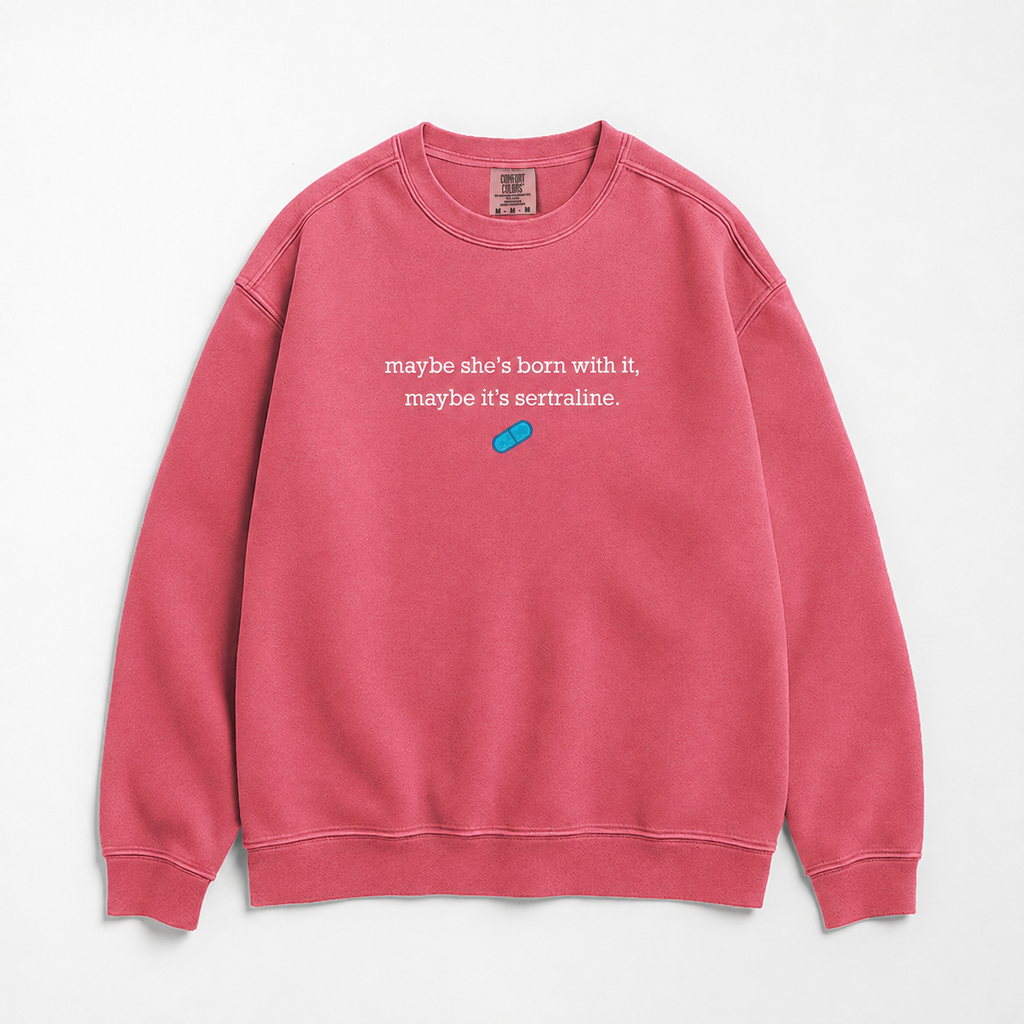 A pink crewneck sweatshirt with white text reading 'maybe she's born with it, maybe it's sertraline.' and a small blue pill graphic on the chest, displayed flat against a light gray background.