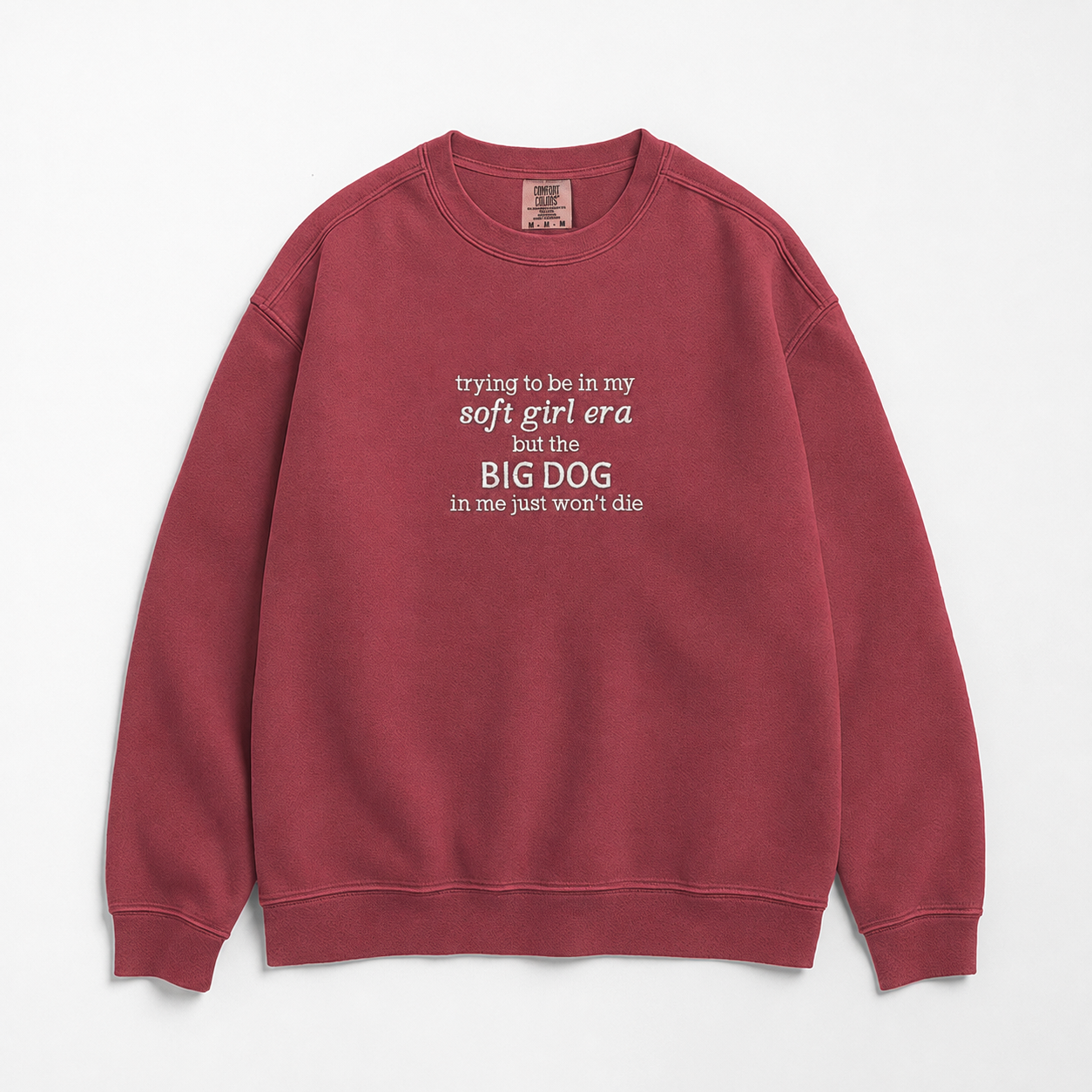 A dusty rose red crewneck sweatshirt with white printed text on the front reading 'trying to be in my soft girl era but the BIG DOG in me just won't die', displayed flat against a light gray background.