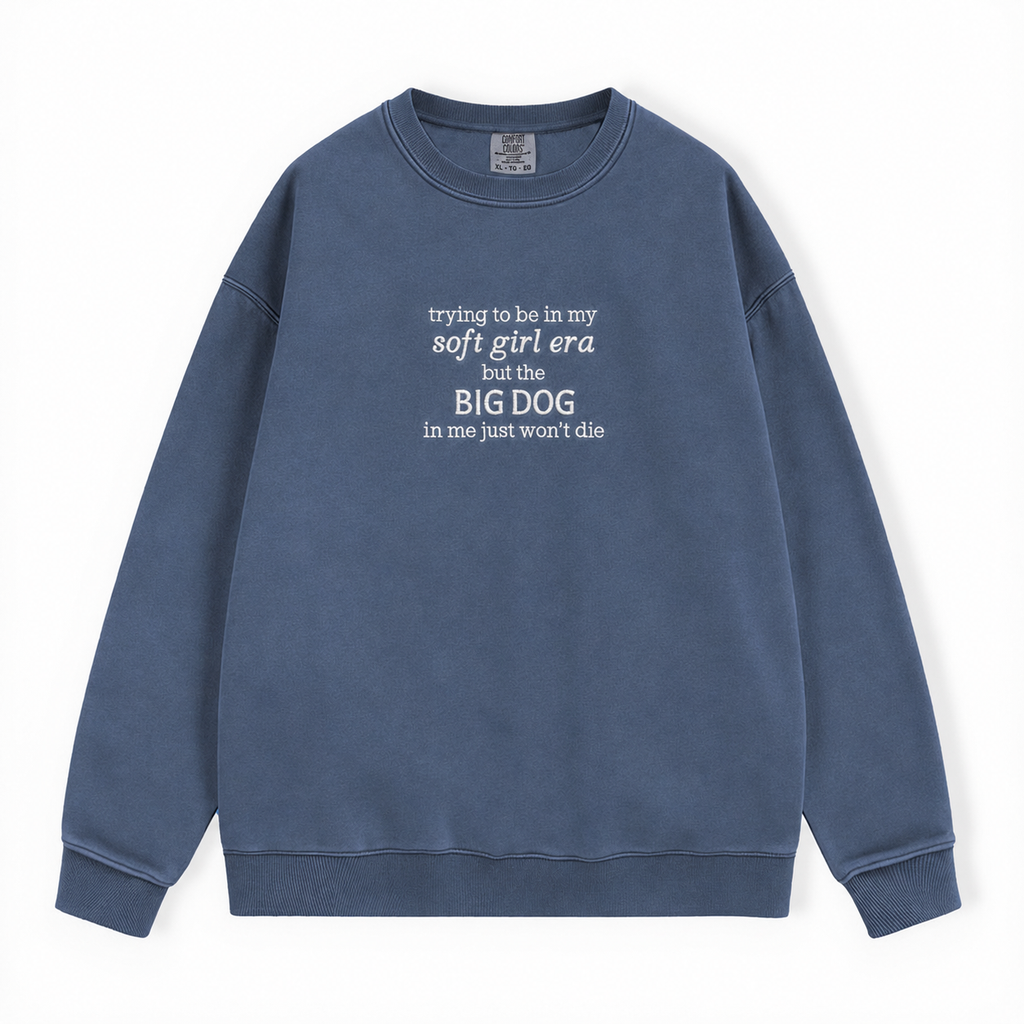 A washed slate blue crewneck sweatshirt with white text on the chest reading 'trying to be in my soft girl era but the BIG DOG in me just won't die', displayed flat against a white background.