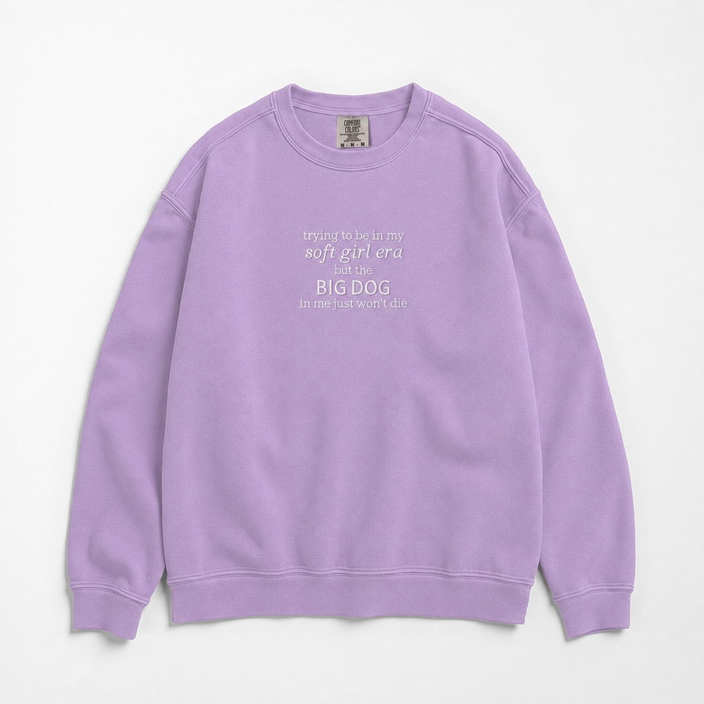 A lavender purple crewneck sweatshirt with a white printed text graphic on the chest reading 'trying to be in my soft girl era before BIG DOG but the big dog in me just won't die', displayed flat against a light gray background.