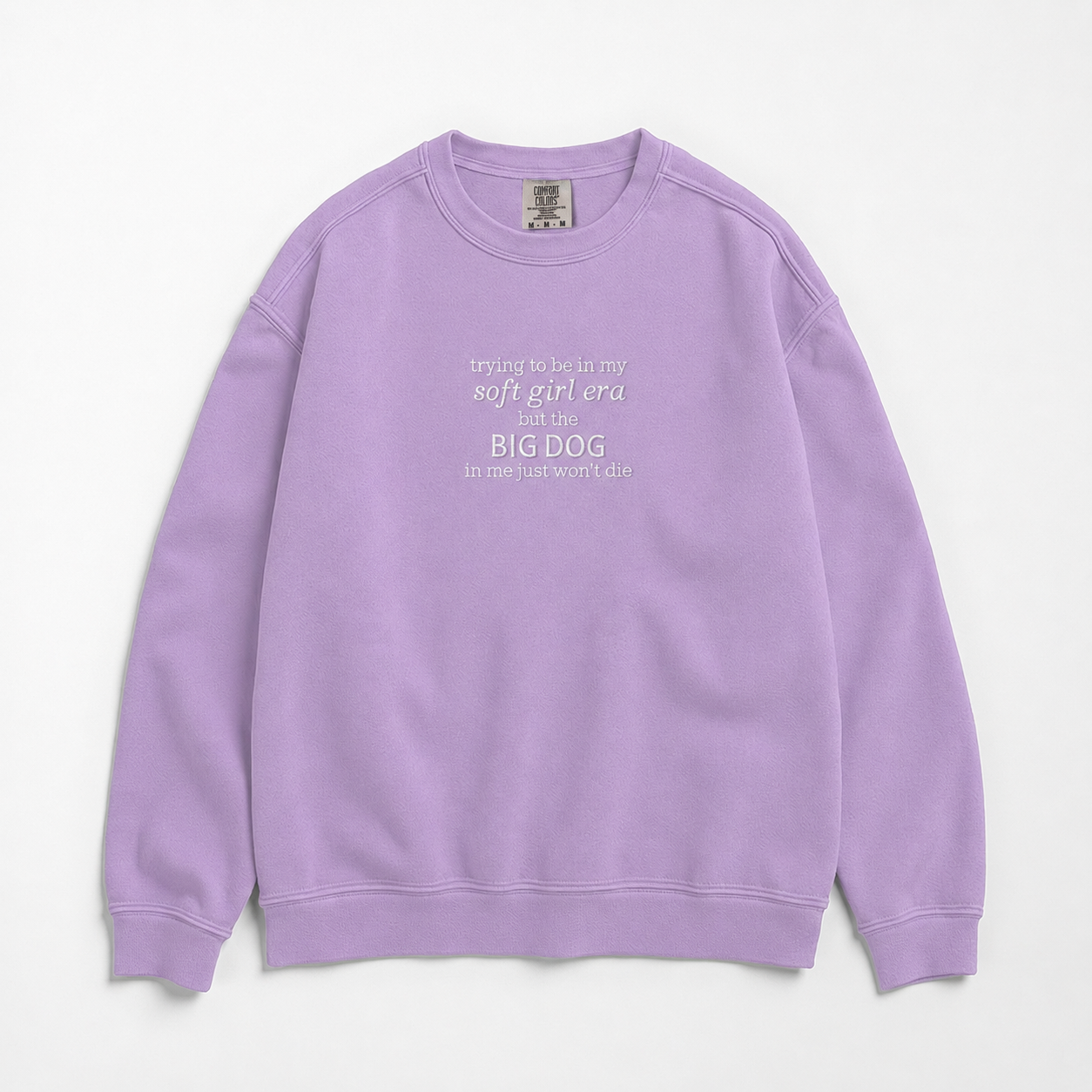 A lavender purple crewneck sweatshirt with a white printed text graphic on the chest reading 'trying to be in my soft girl era before BIG DOG but the big dog in me just won't die', displayed flat against a light gray background.