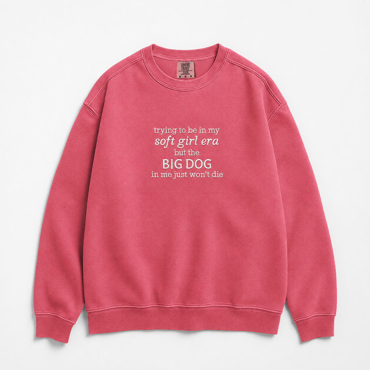 A pink crewneck sweatshirt with white printed text on the chest that reads, 'trying to be in my soft girl era but the BIG DOG in me just won't die', shown against a light gray background.