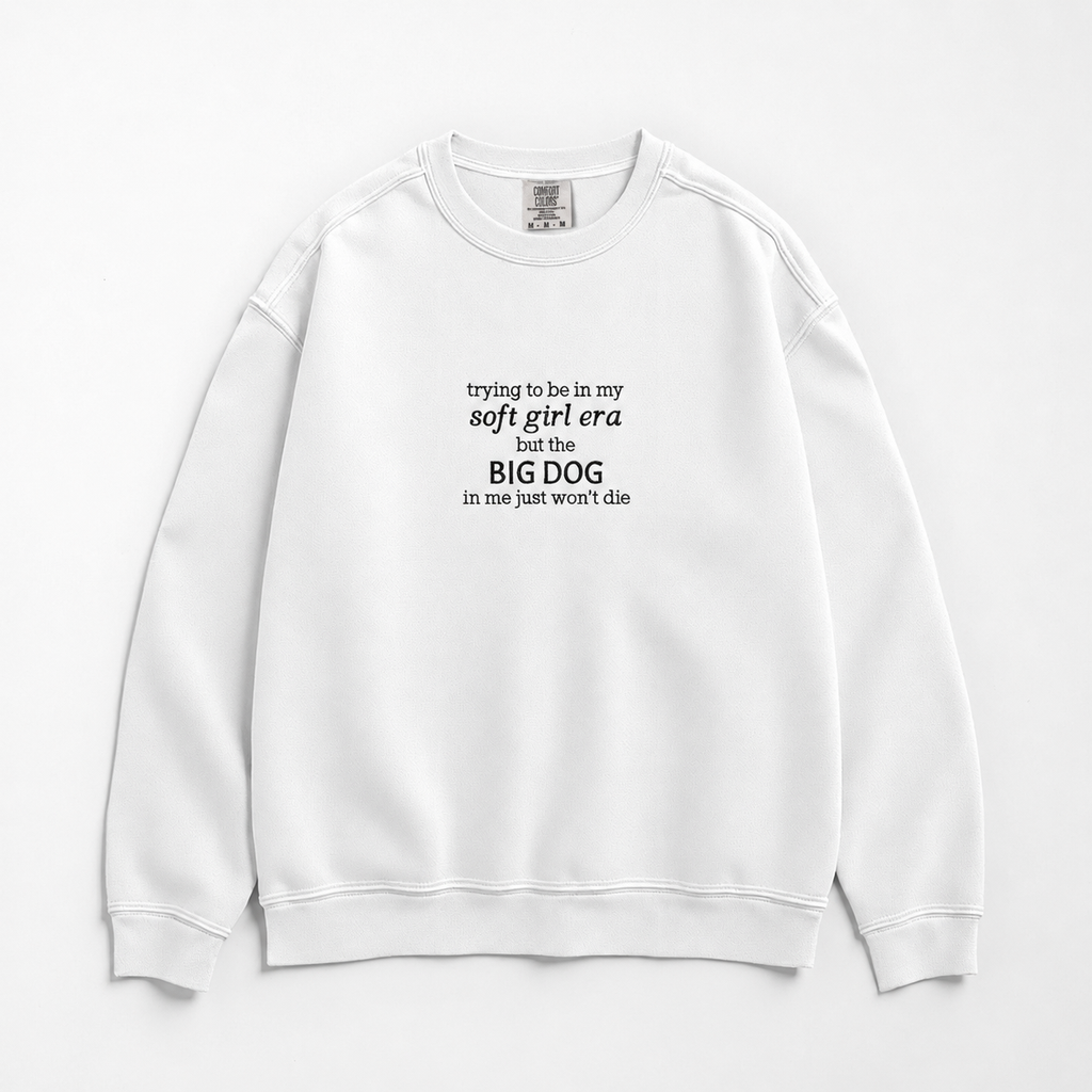 A white crewneck sweatshirt with black printed text on the chest reading 'trying to be in my soft girl era but the BIG DOG in me just won't die', displayed flat against a light gray background.