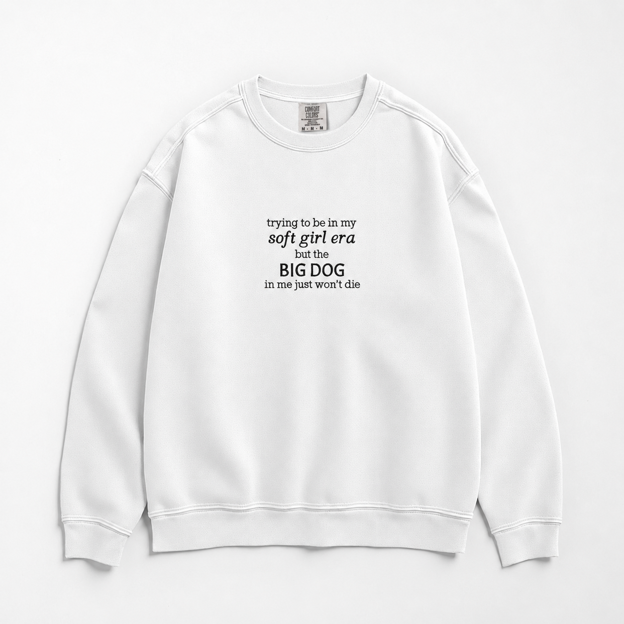 A white crewneck sweatshirt with black printed text on the chest reading 'trying to be in my soft girl era but the BIG DOG in me just won't die', displayed flat against a light gray background.