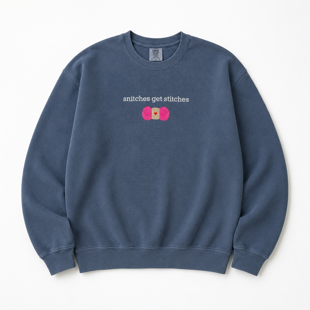 A washed slate blue crewneck sweatshirt featuring a chest embroidered graphic with the text 'snitches get stitches' in white, below a pink skein of yarn.