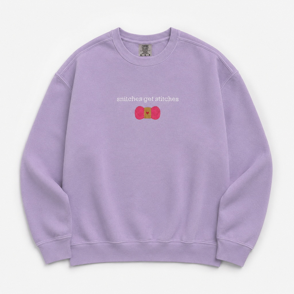 A light purple crewneck sweatshirt featuring a chest embroidered graphic with the text 'snitches get stitches' in white, below a pink skein of yarn.