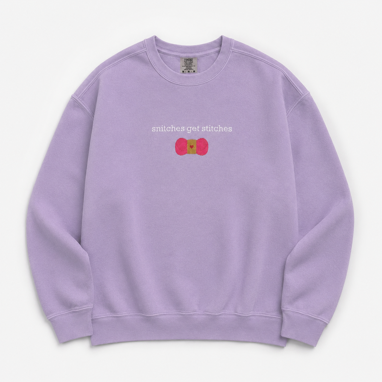 A light purple crewneck sweatshirt featuring a chest embroidered graphic with the text 'snitches get stitches' in white, below a pink skein of yarn.