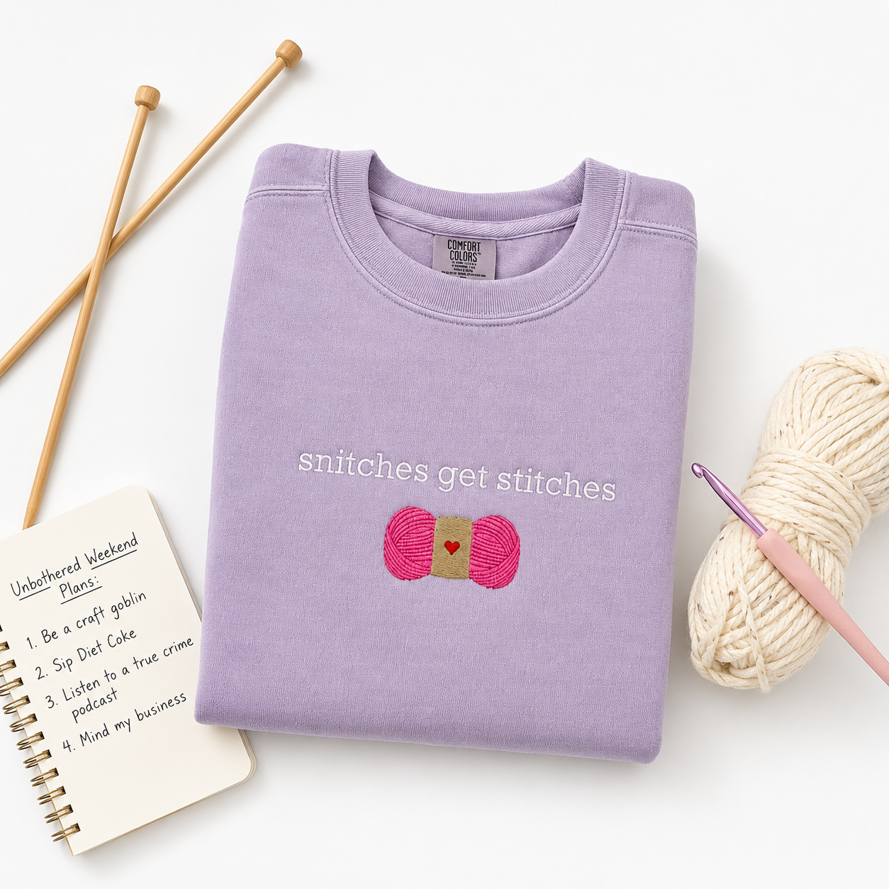A folded lavender crewneck sweatshirt with the text 'snitches get stitches' and a pink yarn graphic embroidered on the front, styled in a flat lay with knitting needles, a crochet hook, a ball of yarn, and a notebook on a white background.