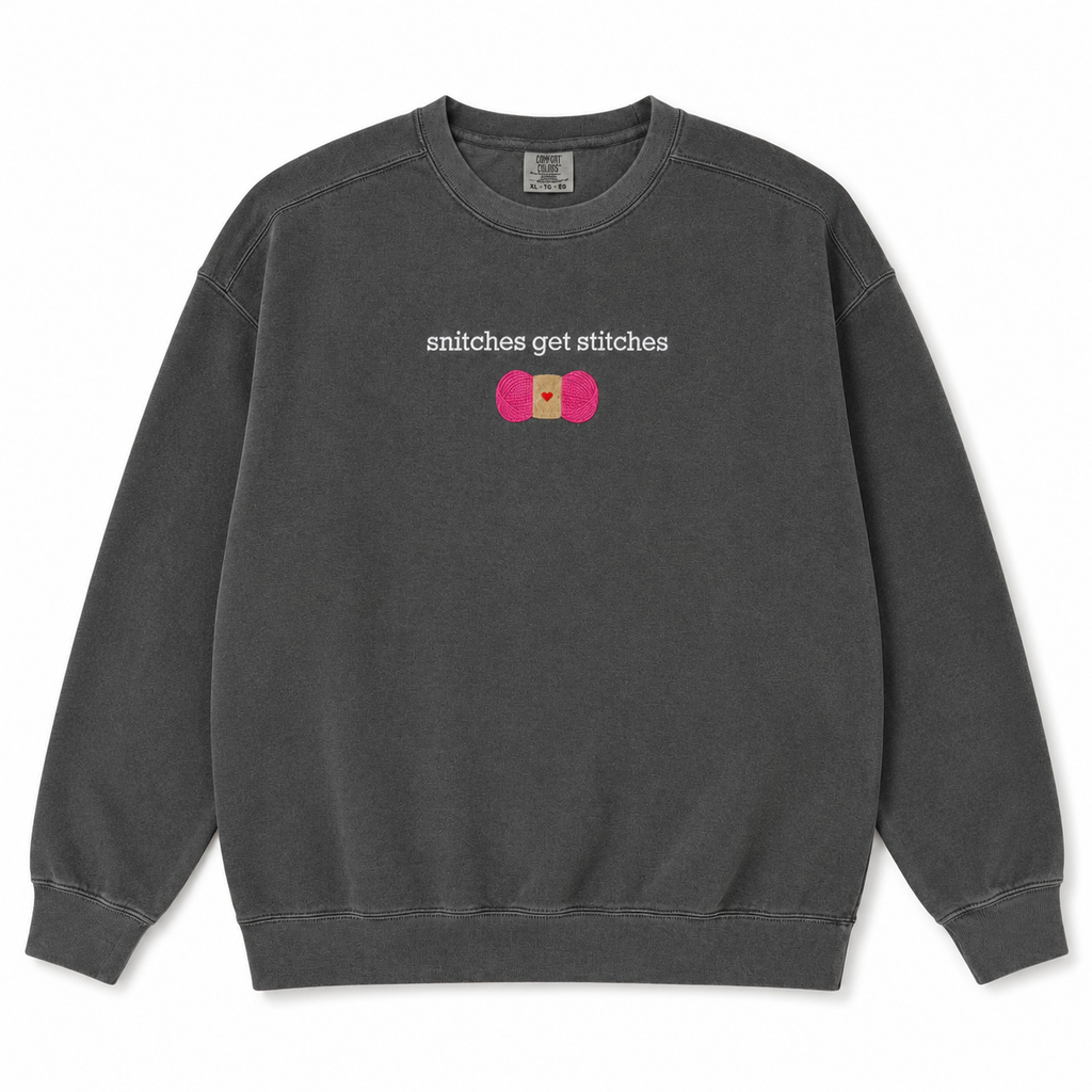 A dark gray crewneck sweatshirt featuring a chest embroidered graphic with the text 'snitches get stitches' in white, below a pink skein of yarn.