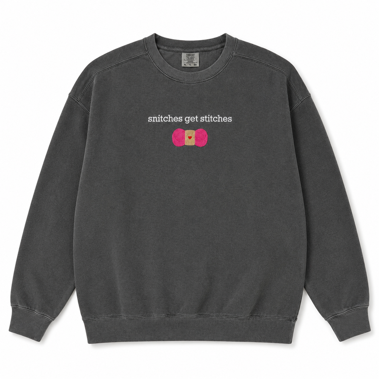 A dark gray crewneck sweatshirt featuring a chest embroidered graphic with the text 'snitches get stitches' in white, below a pink skein of yarn.