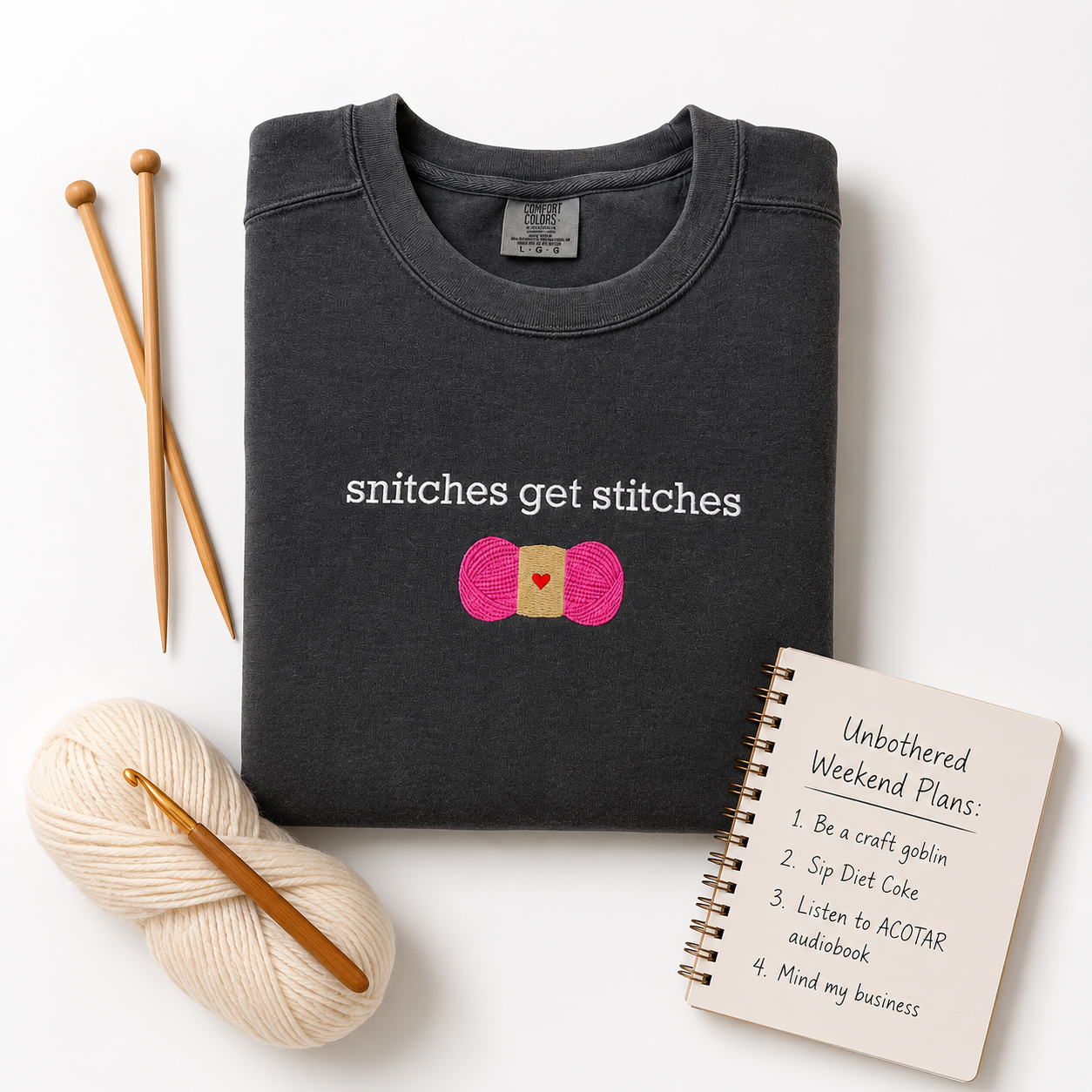 A folded dark charcoal crewneck sweatshirt with the text 'snitches get stitches' and a pink and gold yarn embroidered graphic, styled in a flat lay with knitting needles, cream yarn, a crochet hook, and a spiral notebook on a white background.