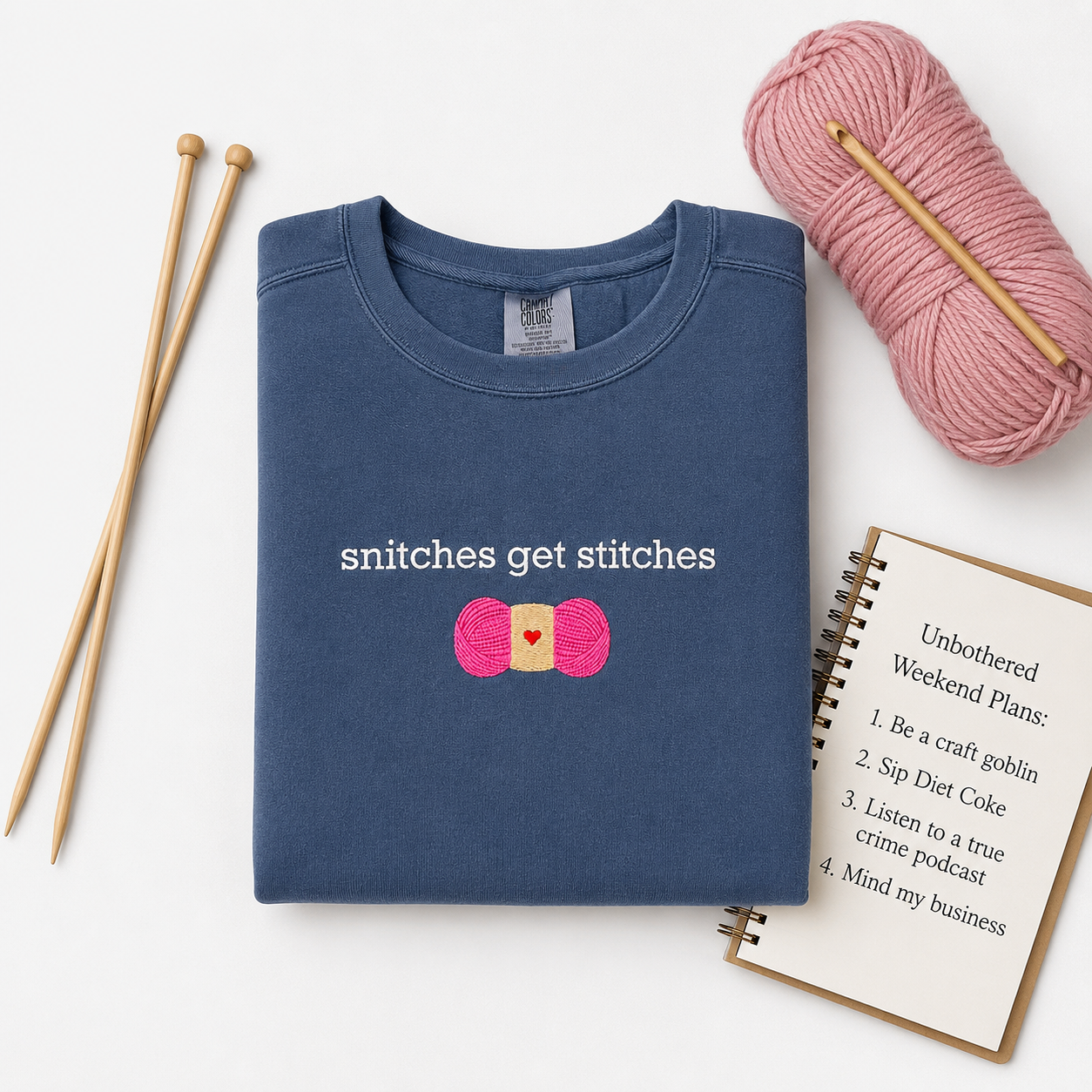 A folded navy blue crewneck sweatshirt with a graphic embroidered as 'snitches get stitches' and a pink yarn ball motif, styled in a flat lay with wooden knitting needles, a pink skein of yarn, and a spiral notebook on a white background.
