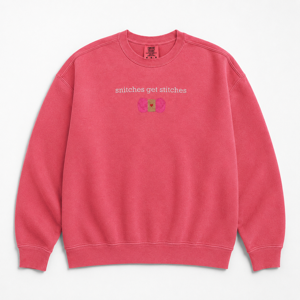 A pink crewneck sweatshirt featuring a chest embroidered graphic with the text 'snitches get stitches' in white, below a pink skein of yarn.